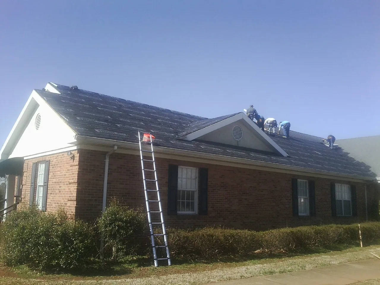 Crew tearing shingles off a brick ranch roof with a ladder set — Metal Roof Repair work by our crew in Flora