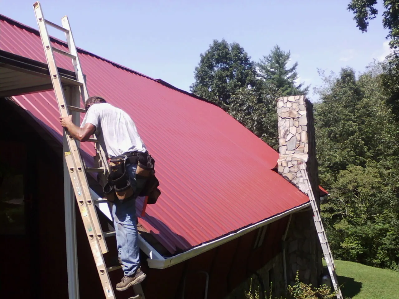 Roofer on a ladder beside a red metal roof and stone chimney — Soffit & Fascia Repair by Roofing Brothers in Flora
